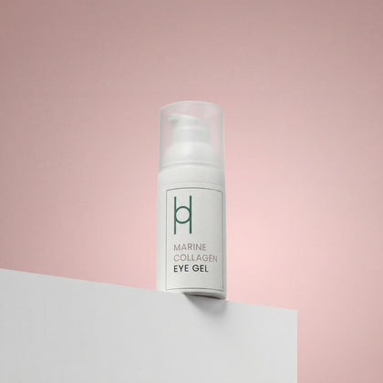 A white pump bottle of Marine Collagen Eye Gel with Hyaluronic Acid, Avocado Oil & Green Tea Extract (30ml) rests on a white surface against a pink background. Minimal green text notes its benefits for dark circles and puffiness, plus a simple logo.