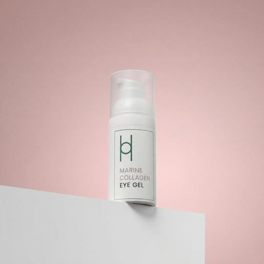 A white pump bottle of Marine Collagen Eye Gel with Hyaluronic Acid, Avocado Oil & Green Tea Extract (30ml) rests on a white surface against a pink background. Minimal green text notes its benefits for dark circles and puffiness, plus a simple logo.