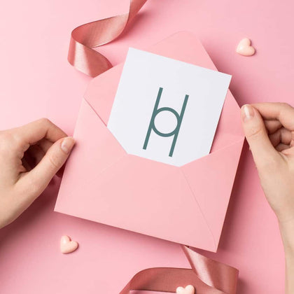 Someone places a Gift me Card, featuring a minimalist geometric design, into a pink envelope. The scene has a pink background with heart-shaped decorations and a satin ribbon.