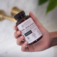 A hand holds a brown bottle labeled Harrogate Organics Company Hair, Skin & Nails Complex and Skincare Bundle, featuring 60 capsules designed to support beauty with a wealth of enriching nutrients. A blurred faucet and white surface are visible in the background, highlighting this essential step in your comprehensive 360-degree skincare routine.