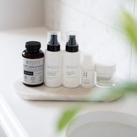 The Hair, Skin & Nails Complex and Skincare Bundle from Harrogate Organics Co.—featuring a supplement bottle, cleanser, toner, serum, and moisturizer enriched with nutrients for complete 360-Degree skincare—is neatly displayed on a marble tray by the sink, with a blurred green plant adding a fresh touch in the foreground.