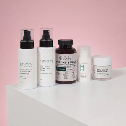 Five products from the Hair, Skin & Nails Complex and Skincare Bundle by Harrogate Organics are displayed in a row on a white surface with a pink background—including cleanser, toner, capsules, serum, and moisturizer—all expertly crafted to deliver enriching nutrients for comprehensive 360-degree skincare.