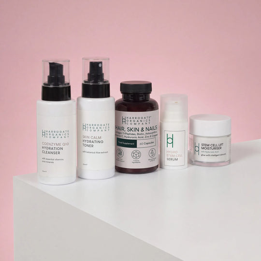 Five products from the Hair, Skin & Nails Complex and Skincare Bundle by Harrogate Organics are displayed in a row on a white surface with a pink background—including cleanser, toner, capsules, serum, and moisturizer—all expertly crafted to deliver enriching nutrients for comprehensive 360-degree skincare.