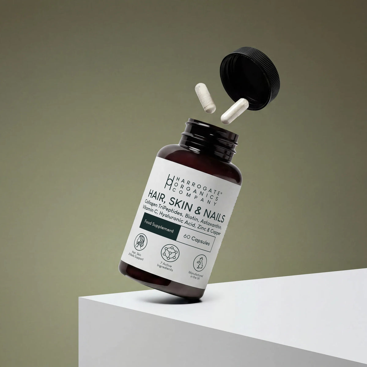 A bottle of Hair, Skin & Nails Complex beauty supplement with two marine collagen-infused capsules floating above the open lid, placed on a white surface against a muted background.