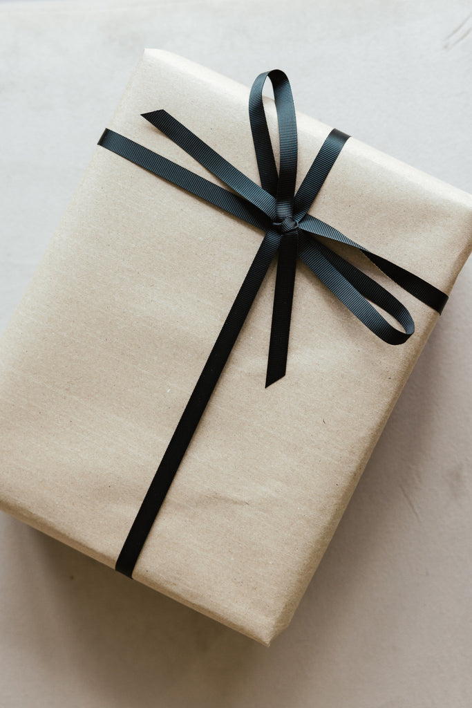 Gift Wrap: A rectangular box covered in plain brown paper and finished with a neat black ribbon bow, placed on a light surface—simple wrapping that adds a special touch.