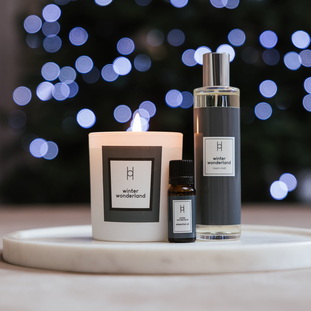 A lit candle, a small essential oil blend, and a tall spray bottle from the Deluxe Home Collection - Winter Wonderland rest on a marble tray, with twinkling festive lights softly blurred in the background.