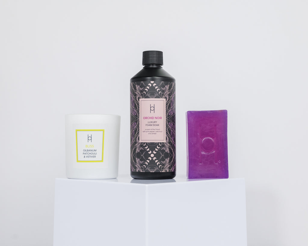 The Indulgent Bath Trio, featuring a white candle, black Orchid Noir Foam Soak bottle, and purple soap bar, is displayed on a white pedestal against a minimalist backdrop for a spa-like experience.