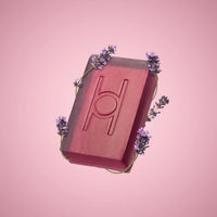 The Lavender Fields - Luxe Soap is a rectangular vegan bar in pink, featuring an engraved geometric design and wrapped with sprigs of purple lavender, presented against a soft pink background.