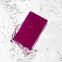 The Lavender Fields - Luxe Soap, a rectangular translucent purple bar with an embossed design, splashes into clear water, sending dynamic ripples and droplets outward.