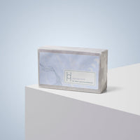 A moisturising Lavender Fields - Luxe Soap bar in translucent packaging rests on a white geometric surface with a light blue gradient background, promising clean and nourished skin.