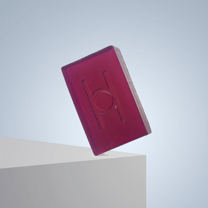 A translucent, rectangular magenta bar of Lavender Fields - Luxe Soap with an embossed “H” and circle design balances on a white edge against a light blue gradient background.