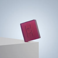 The Lavender Fields - Luxe Soap, a translucent purple bar with an embossed “H” logo, stands on its edge on a white surface set against a light blue gradient background.