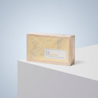 A rectangular bar of Luxe Soap in yellow packaging, labeled "Lemon Grove" with a subtle fish illustration, sits on a white surface against a light blue background, hinting at the refreshing nature of this moisturizing soap.
