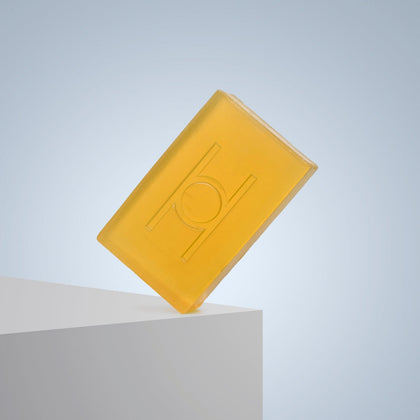 The Lemon Grove - Luxe Soap, a translucent yellow vegan bar with a raised geometric pattern, stands upright on the edge of a white surface against a light blue gradient background.