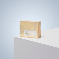 A yellow rectangular bar of Lemon Grove - Luxe Soap sits on a white surface with a light blue gradient background. The moisturising soap’s packaging features subtle floral patterns and promises clean, nourished skin.