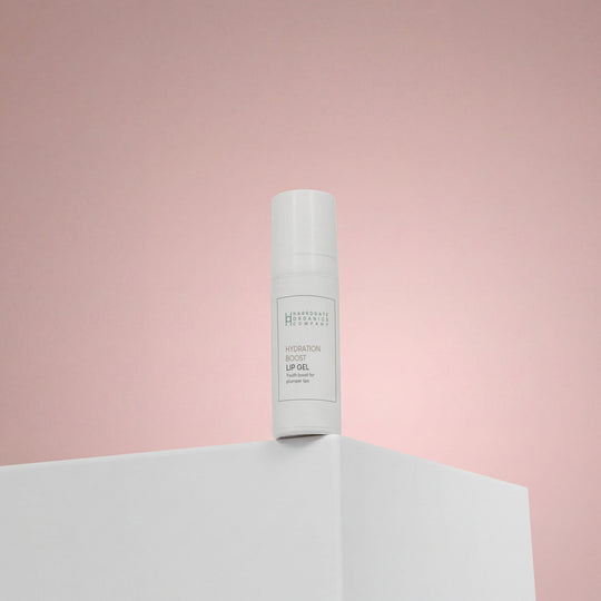 A white bottle labeled Hydration Boost Lip Gel | Fuller, Hydrated Lips | 10ml stands upright on a white surface with a soft pink gradient backdrop.