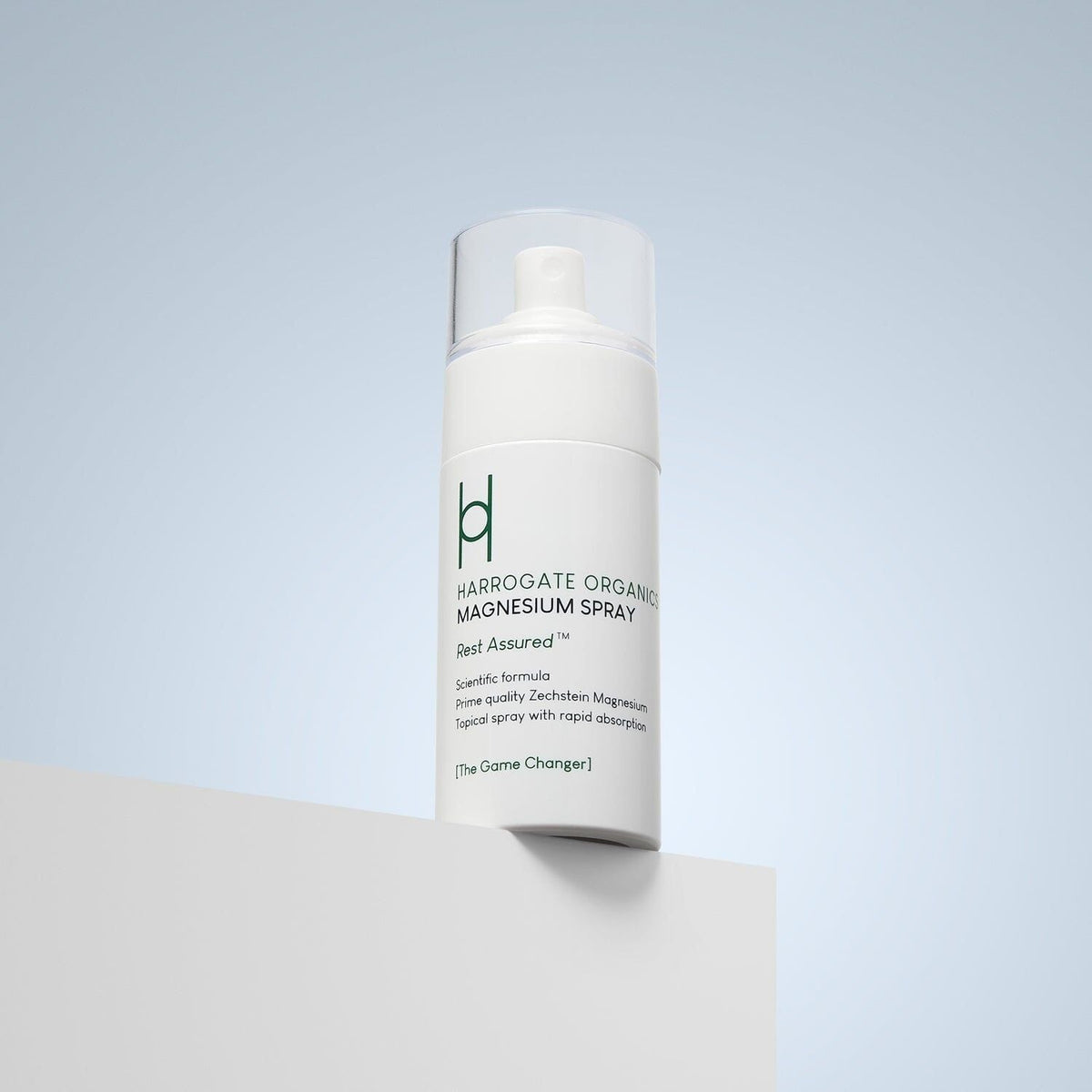 A white bottle of Harrogate Organics Magnesium Spray - 100ml sits on a white surface with a pale blue background. The label reads "Rest Assured" and highlights organic wellness and rapid absorption benefits.