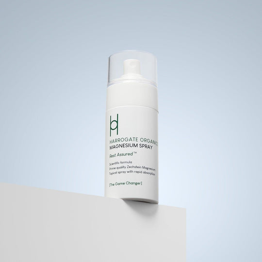 A white bottle of Harrogate Organics Magnesium Spray - 100ml sits on a white surface with a pale blue background. The label reads "Rest Assured" and highlights organic wellness and rapid absorption benefits.