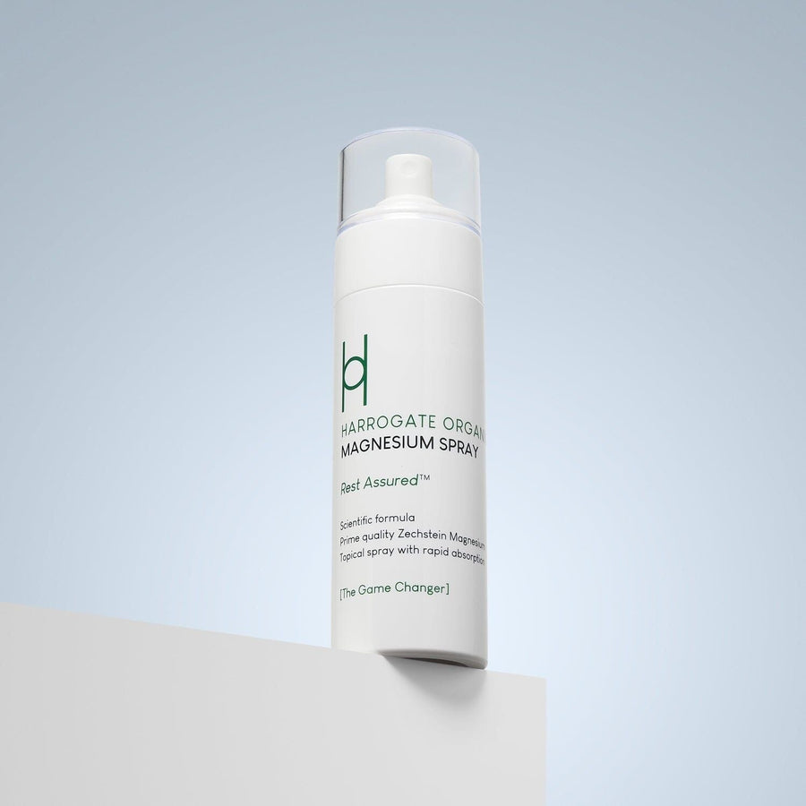 A white bottle of Harrogate Organics Magnesium Spray - 150ml with a clear cap sits on a white surface against a light blue background. The label displays product details and the phrase "Rest Assured.
