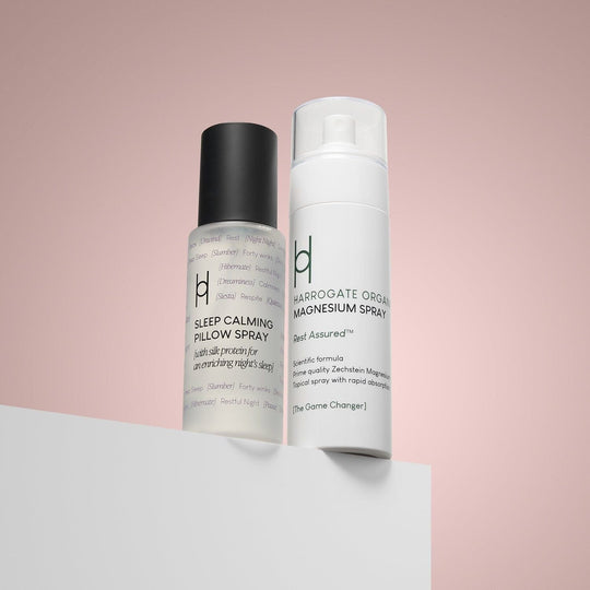 Two bottles—Magnesium Spray and Pillow Mist—stand side by side on a white surface with a pink background, ideal for rest and relaxation.
