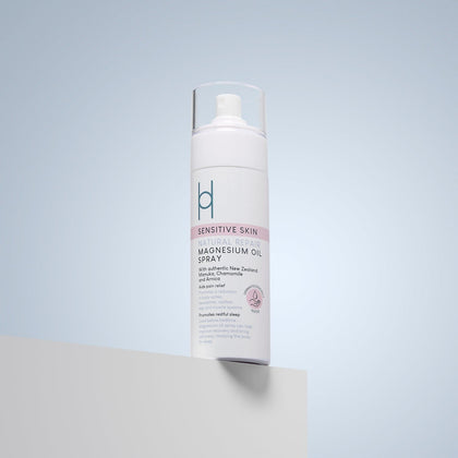 A white bottle of sensitive skin natural repair magnesium oil spray stands on a light gray surface against a pale blue background. The label features blue and pink text and a logo near the top.