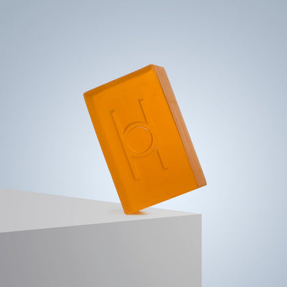 A rectangular orange Fresh Mandarin - Luxe Soap bar with an embossed design stands on its edge atop a white surface against a light blue background—this vegan, moisturizing soap adds vibrancy to your routine.