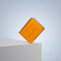 A translucent orange Fresh Mandarin - Luxe Soap bar with circular and line designs balances on a white edge against a light blue background, showcasing the vibrant allure of this vegan soap.