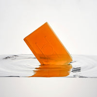 A translucent amber bar of Fresh Mandarin - Luxe Soap embossed with an H is partially submerged in clear water, creating gentle ripples against a white background—perfect for moisturising and radiant skin.