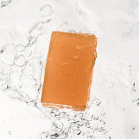 A translucent orange Fresh Mandarin - Luxe Soap bar with an embossed design is partially submerged in clear water with splashes and bubbles, offering a refreshing, moisturising vegan soap experience.