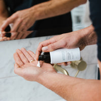 A person dispenses cleanser from the Ultimate Men's Skincare Set’s black and white bottle into their hand over a marble countertop and brass faucet, starting their men’s skincare regimen.