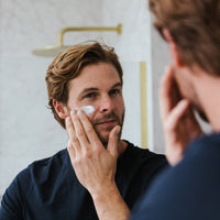 A man with light brown hair applies the Ultimate Men's Skincare Set | Complete 4-Piece Collection to his cheek while looking in the bathroom mirror, demonstrating his commitment to a comprehensive men's skincare routine.