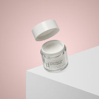 A glass jar from the Women's Skin Reset Gift Set | 4-Piece Anti-Ageing Collection sits on a white surface with its lid floating above, minimal branding visible, against a pink gradient background to highlight its advanced benefits.