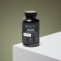 A dark bottle of Harrogate Organics Company NMN, Magnesium, Zinc & Collagen Supplement sits on a white and gray surface with a gray-green background. The label highlights 700mg NMN per serving, collagen for healthy skin, magnesium for energy, zinc, and contains 60 capsules for a 30-day supply.