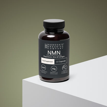 A dark bottle of Harrogate Organics Company NMN, Magnesium, Zinc & Collagen Supplement sits on a white and gray surface with a gray-green background. The label highlights 700mg NMN per serving, collagen for healthy skin, magnesium for energy, zinc, and contains 60 capsules for a 30-day supply.