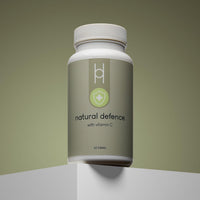 A white bottle of Natural Defence with Vitamin C, featuring a shield and cross icon, sits on a white pedestal against a gradient olive background; the wellness supplement contains 60 tablets and is made with natural ingredients.