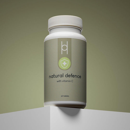 A white bottle of Natural Defence with Vitamin C, featuring a shield and cross icon, sits on a white pedestal against a gradient olive background; the wellness supplement contains 60 tablets and is made with natural ingredients.