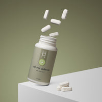 A bottle of Natural Defence with Vitamin C lies on its side on a white surface, white tablets spilling out and floating above. The background is green.