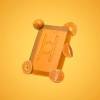 A rectangular orange Fresh Mandarin - Luxe Soap bar with a carved logo sits amid whole and halved oranges on a yellow-orange gradient background. This vegan soap delivers a burst of freshness to your daily routine.