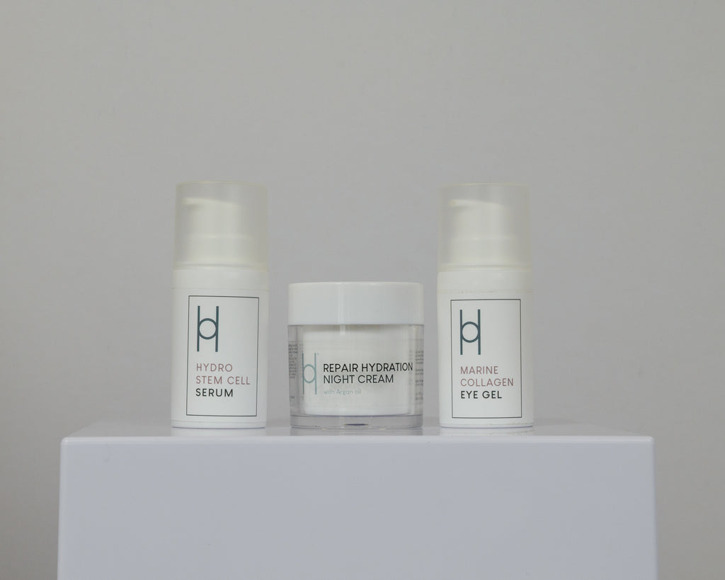 The Overnight Repair Trio—Night Cream, Eye Gel, and Hydro Stem Cell Serum in sleek white containers with minimalist labels—rests on a white platform against a plain backdrop.