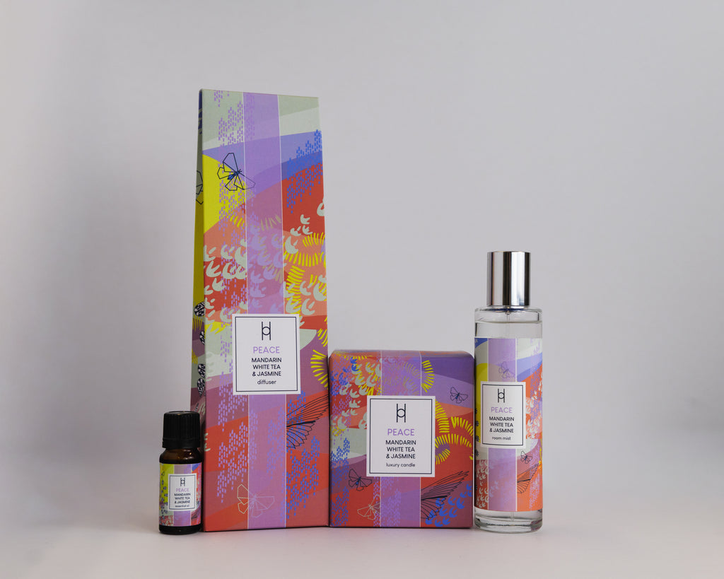 Four products in colorful, abstract-patterned packaging sit on a white background: an essential oil bottle, the Deluxe Home Collection - Peace home fragrance box, a larger rectangular box, and a clear reed diffuser bottle with a silver cap.