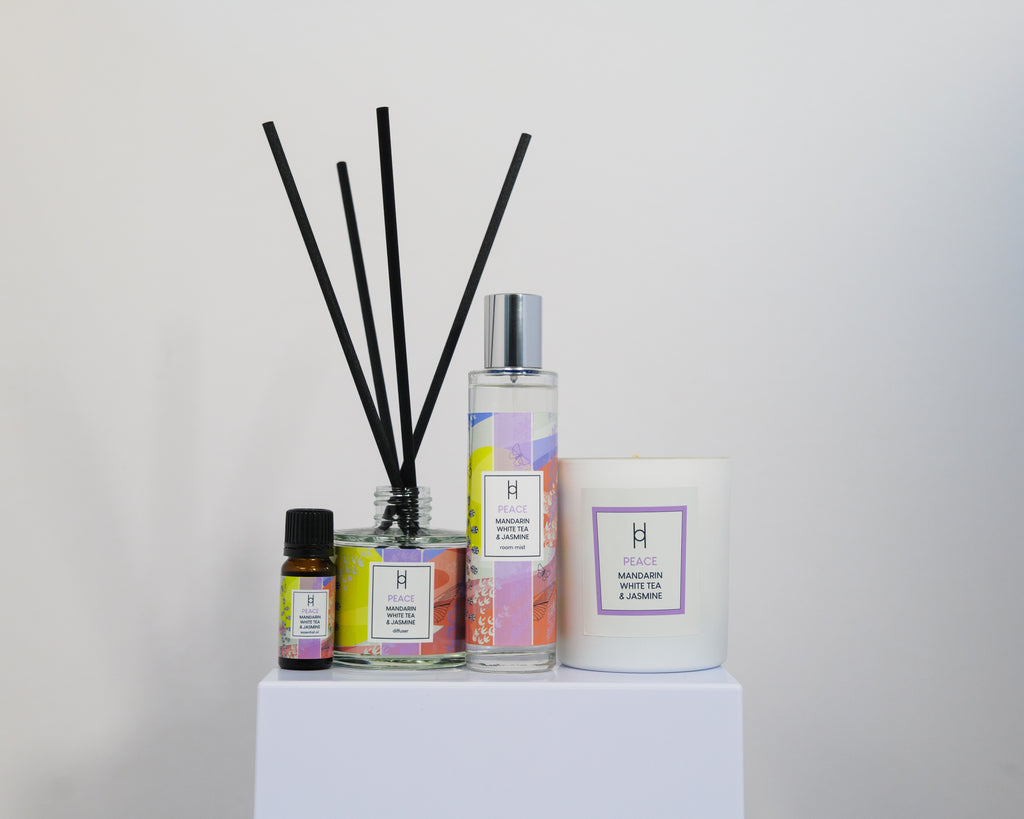 The Deluxe Home Collection - Peace features mandarin and jasmine scent in a reed diffuser with black reeds, a spray bottle, essential oil, and a white candle, all elegantly arranged on a white pedestal against a plain background.
