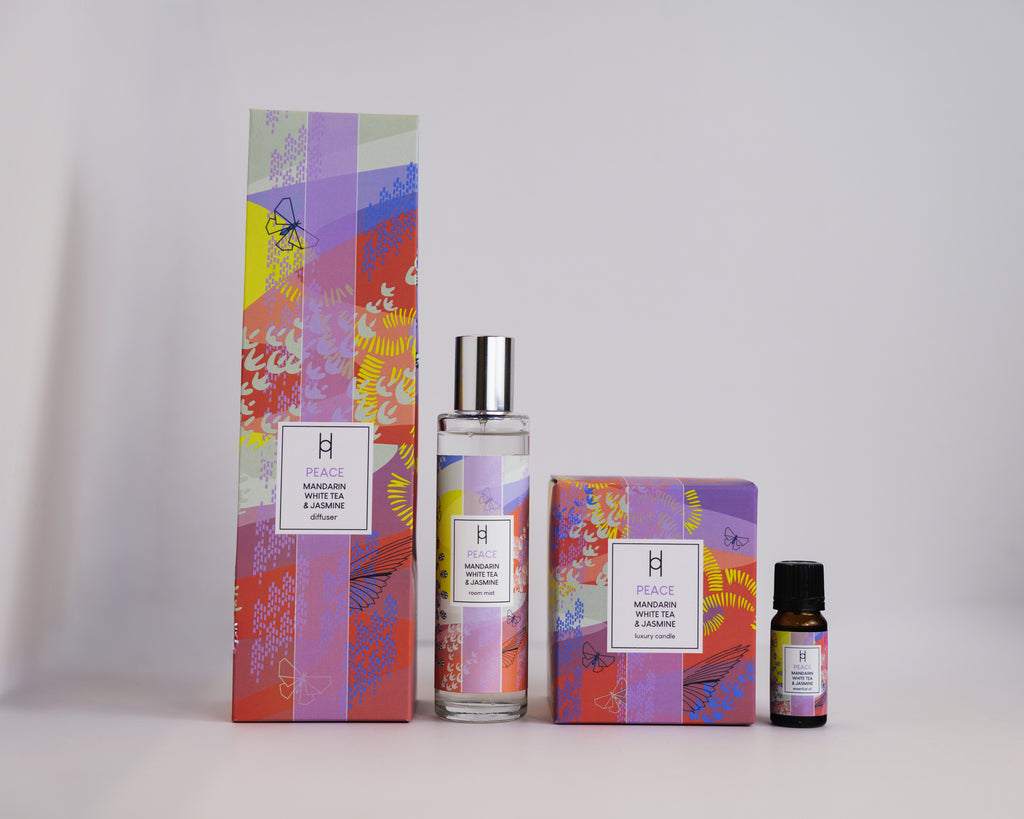 The Deluxe Home Collection - Peace features colorful, abstract packaging on a tall box, spray bottle, square box, and small essential oil bottle, all set against a plain light background.