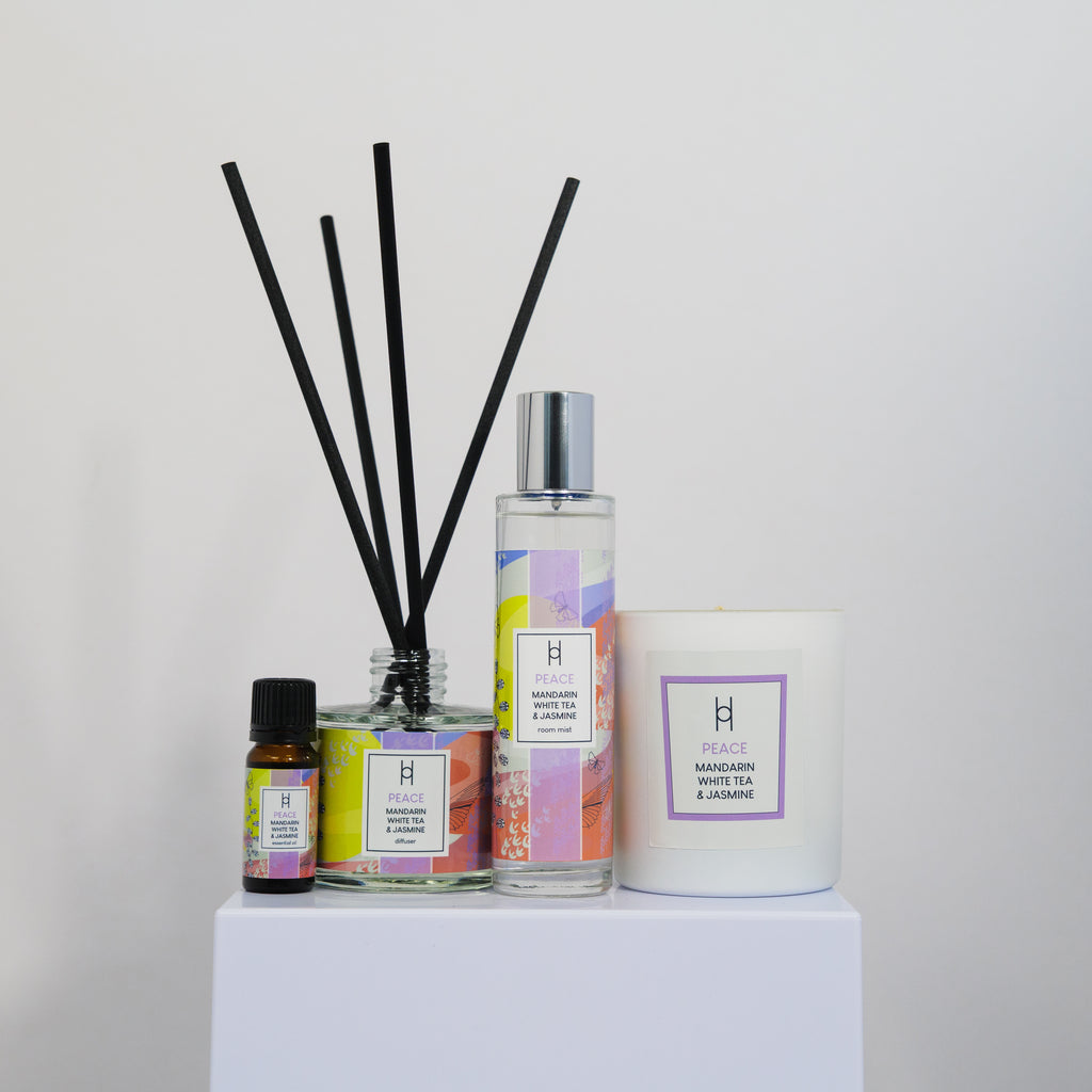 The Deluxe Home Collection - Bliss is displayed on a white pedestal, featuring four home fragrance items: an essential oil bottle, reed diffuser, spray bottle, and a white candle against a plain white background.