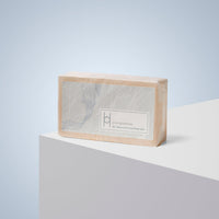 A rectangular bar of Pure Goodness Soap - Unscented, wrapped in beige paper with a minimalist label, rests on a white surface against a light blue background.