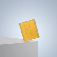 The translucent yellow Pure Goodness Soap - Unscented, embossed with an H, rests on a white rectangular surface against a pale blue background.