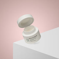 A white jar of Rescue Balm with its lid open is displayed on the edge of a white surface, set against a soft pink gradient background. The label includes green text and minimal design elements.