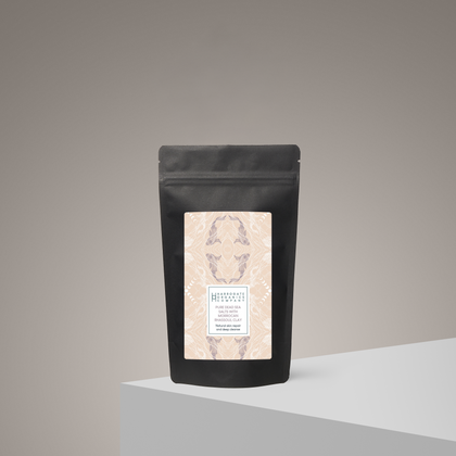 A black resealable bag with a decorative beige and pink label stands upright on a light gray surface. The label features abstract patterns and highlights Pure Dead Sea Salts With Moroccan Rhassoul Clay, a unique spa blend.