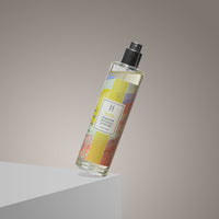 A transparent spray bottle with a vibrant Bliss Room Mist | Ylang Ylang Aromatherapy Spray | 100ml label rests at an angle on a light gray surface, set against a neutral background.