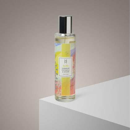 A glass bottle of Bliss Room Mist | Ylang Ylang Aromatherapy Spray, 100ml, with a black cap and colorful abstract design, sits on a white platform against a neutral background.