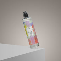 A clear spray bottle with a pastel label reading Peace Room Mist | Lemon, Mandarin Aromatherapy Spray rests at an angle on a white platform against a neutral background, highlighting this all-natural room mist and its calming citrus scent.